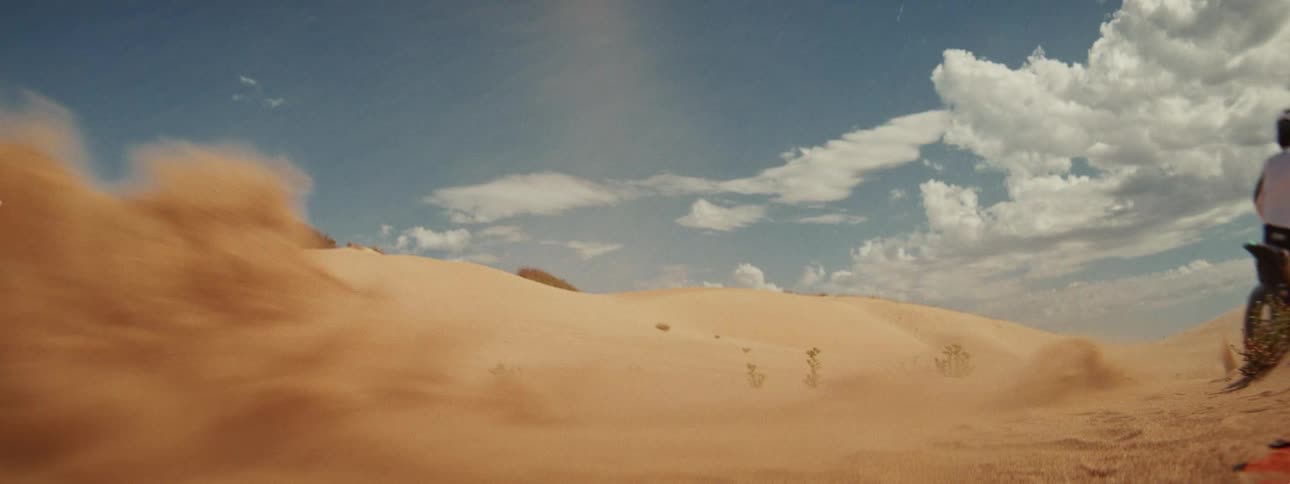 A motocross rider accelerates on a dirt bike in slow motion, kicking up a large cloud of sand as they speed across desert dunes
