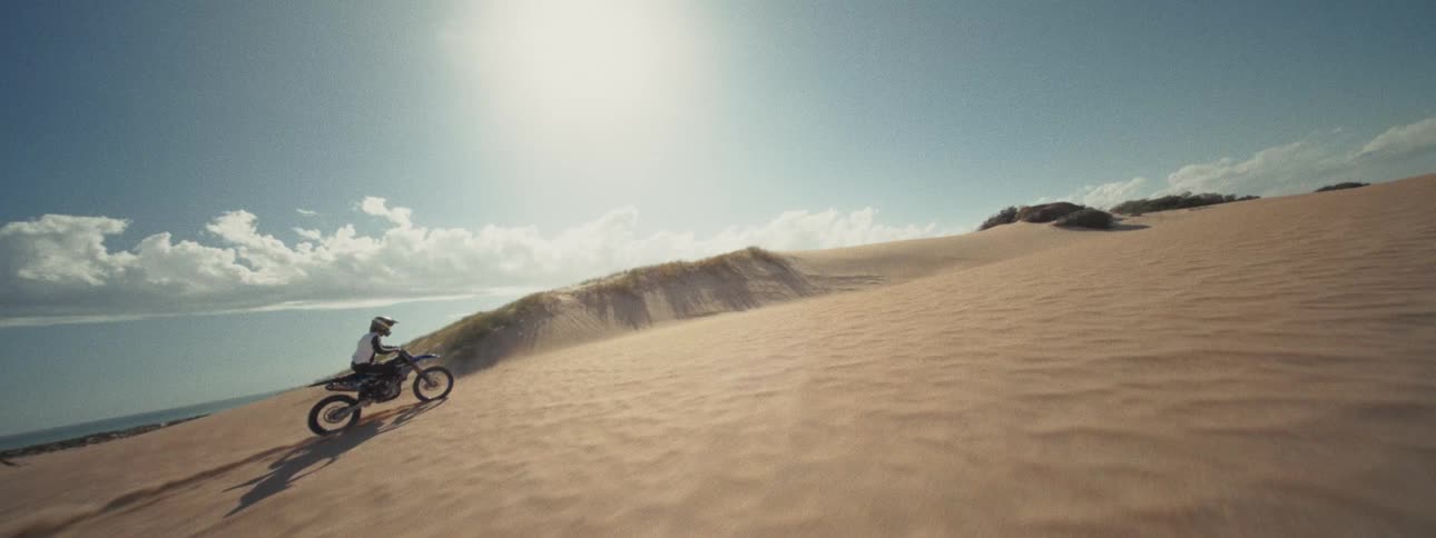 A dynamic aerial shot tracks a motocross rider speeding up and carving through vast, sunny sand dunes