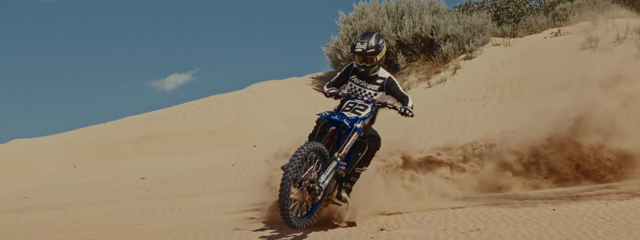 A professional motocross racer wearing full gear powerfully corners on a sand dune, kicking up sand in dramatic slow motion