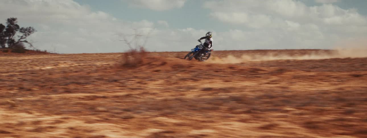 A motocross rider kicks up a huge cloud of dust while speeding through a barren, dry landscape in slow motion