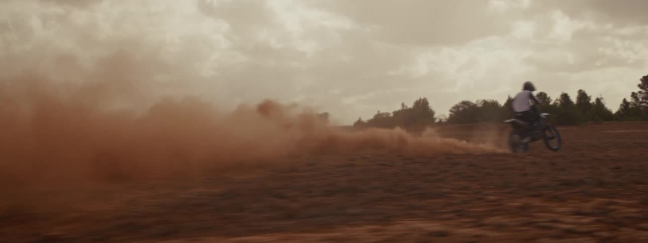 A motocross rider speeds across a dry, dusty landscape, kicking up a massive cloud of red dirt in slow motion