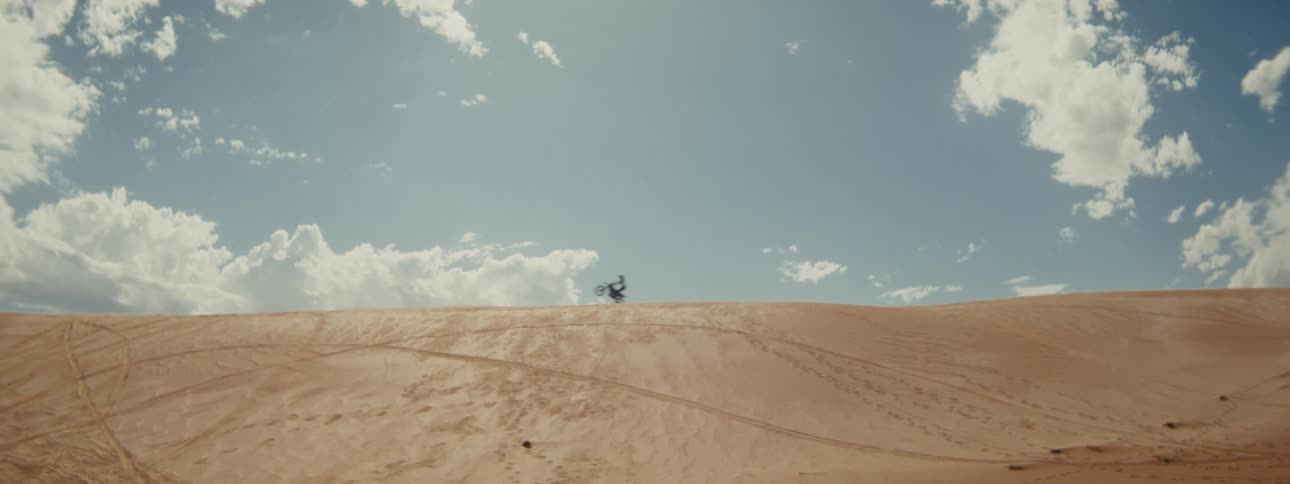 A motorcyclist rides in slow motion across the top of a large sand dune under a partly cloudy blue sky