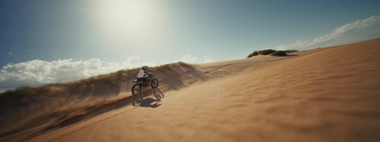 A thrilling FPV drone shot tracks a dirt bike rider as they race across golden sand dunes under a clear blue sky