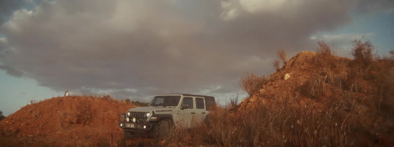 A motocross rider in slow motion jumps over a dirt mound with a rugged off-road Jeep parked in the foreground