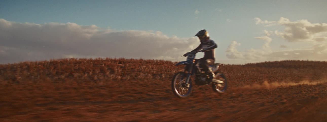 Cinematic slow motion tracking shot of a motocross racer riding through a dusty field and doing a wheelie at sunset