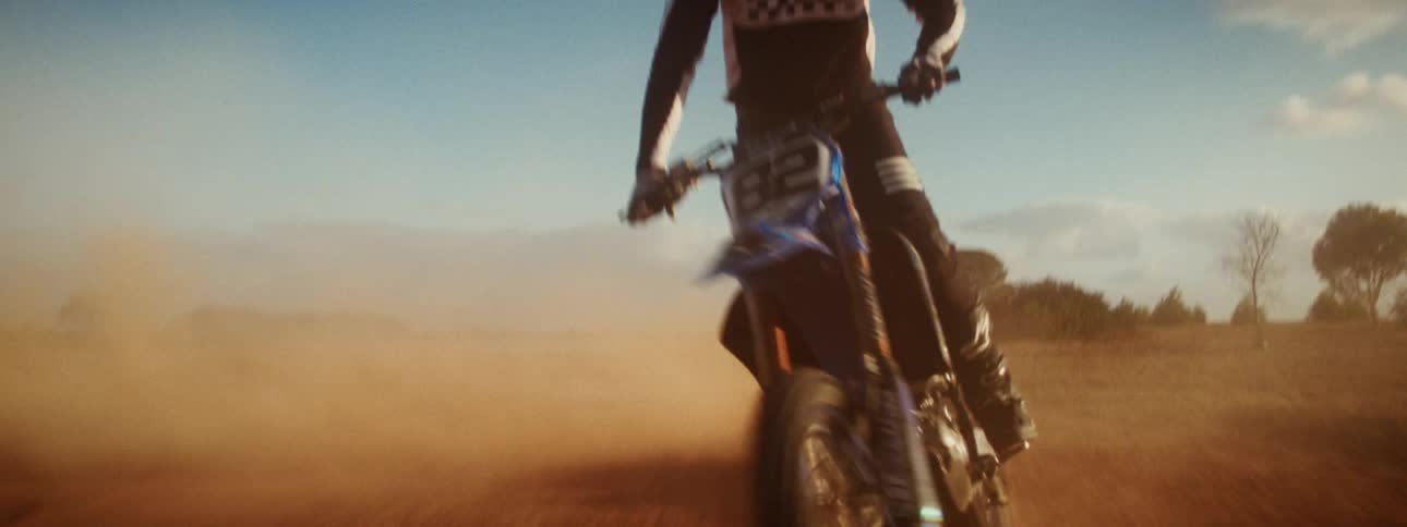 Slow motion footage shows a professional motocross rider doing a wheelie and speeding down a dusty track during golden hour