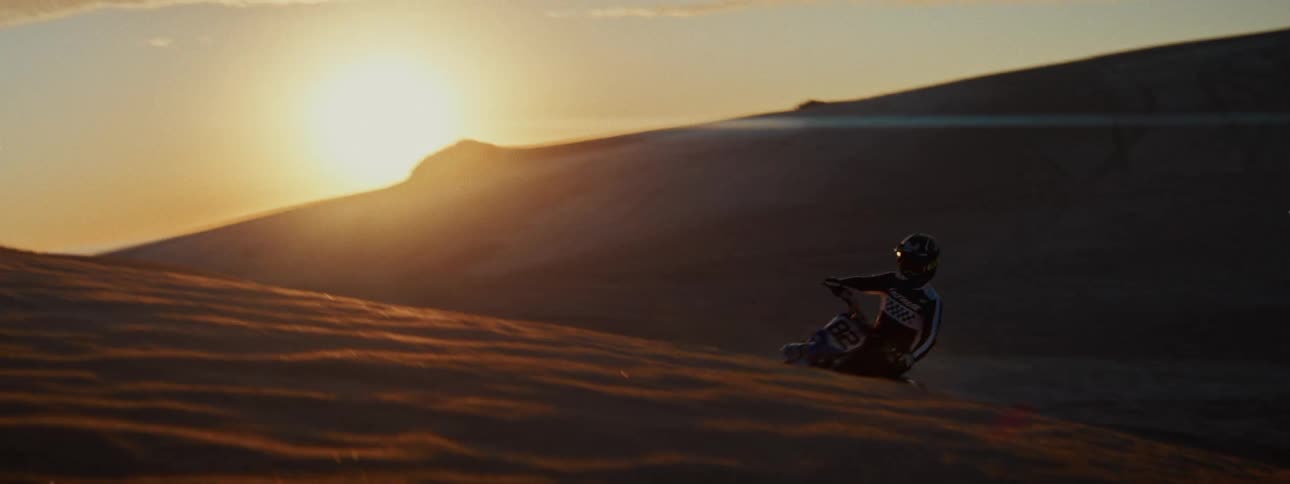 In slow motion, a motocross rider carves a turn on a sand dune, kicking up sand against the golden sunset before falling