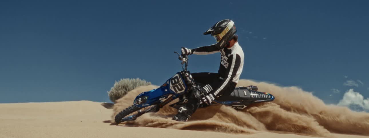 A motocross rider carves a sharp turn through a sand dune, kicking up a dramatic spray of sand in slow motion