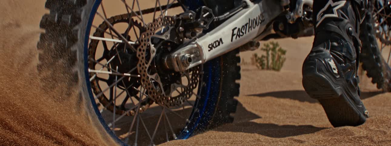 A motocross bike's back tire spins in slow motion, digging into the sand and kicking up a plume of dust