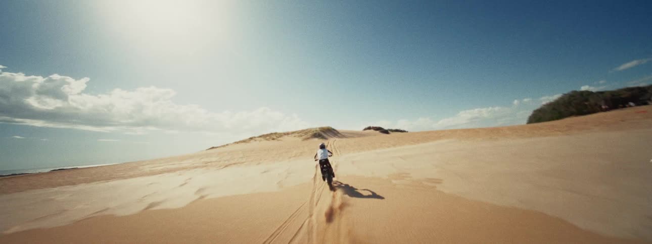An exhilarating aerial shot tracks a lone dirt bike rider carving through golden sand dunes next to the sea