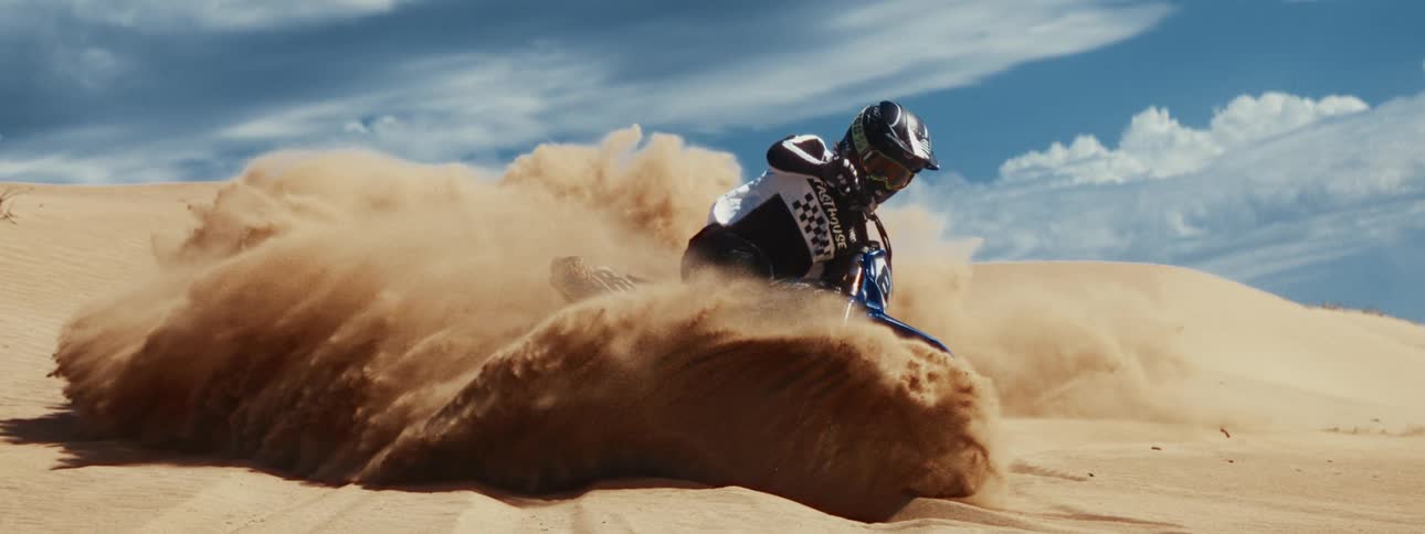 A professional motocross racer skillfully carves through a sand dune, kicking up a massive wave of sand in dramatic slow motion