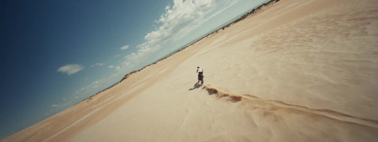 Aerial drone follows a dirt bike rider speeding across expansive desert sand dunes towards the ocean