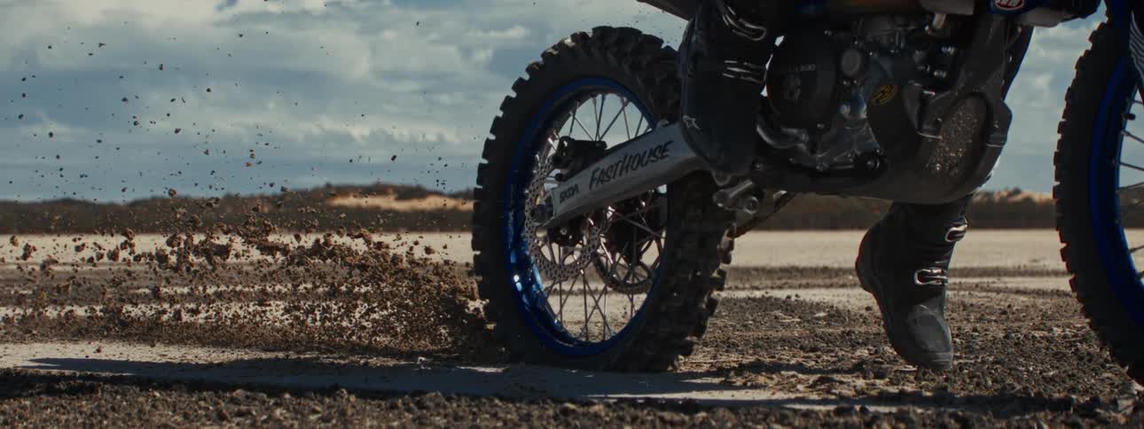 In cinematic slow motion, a dirt bike's rear tire digs into the ground, kicking up a spray of dirt and sand
