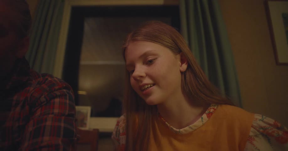 An engaged teenage girl with long hair smiles while having a conversation in a warmly lit room at night