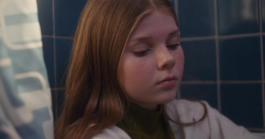 A sad young girl with red hair and freckles sits alone in a bathroom, lost in thought
