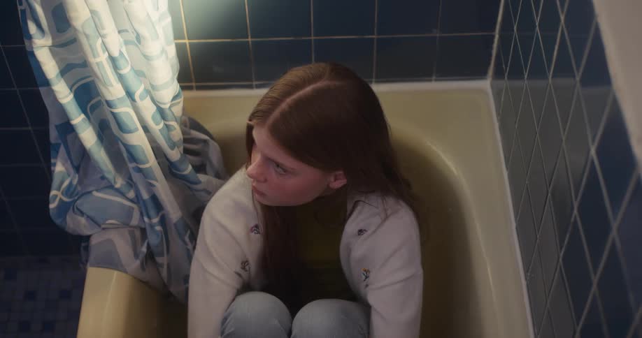 Young woman sits curled up in a bathtub in a blue tiled bathroom with a quiet, pensive mood