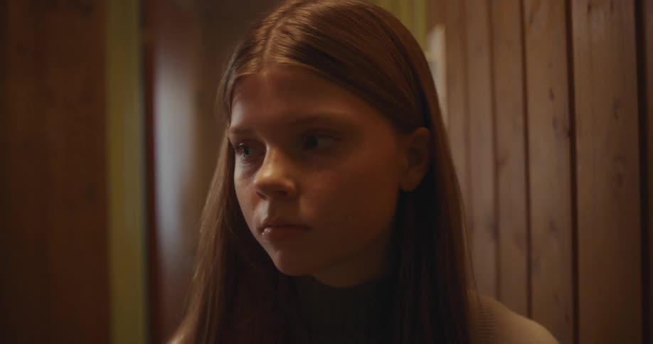 A solemn young girl peeks out from behind a wall and looks around with a worried expression in a dimly lit, moody hallway