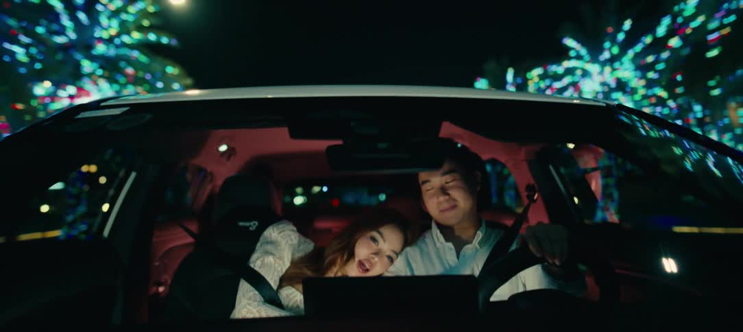 A loving Asian couple enjoys a romantic nighttime drive, holding hands as they pass festive city lights
