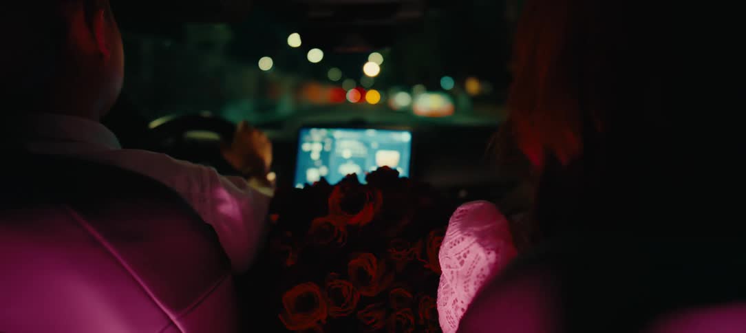 A romantic couple drives through the city at night with a large bouquet of red roses, viewed from the back seat