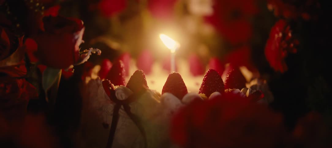A single burning candle illuminates a beautiful strawberry birthday cake surrounded by red roses in a warm, intimate setting