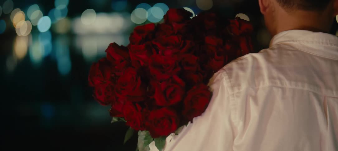 Close up from behind of a man holding a large bouquet of red roses at night with city lights in the background