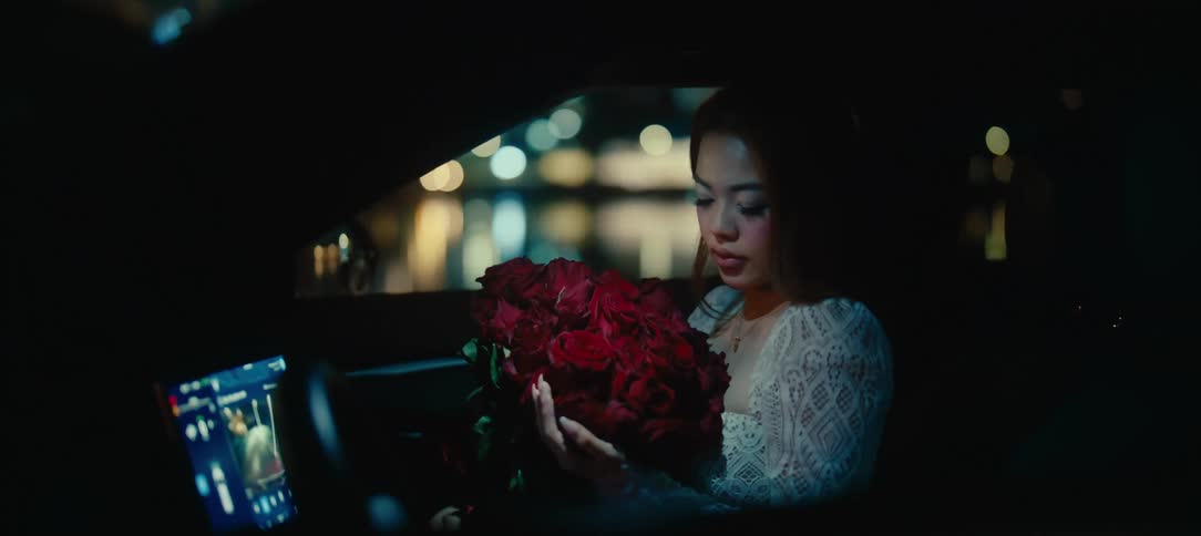 A beautiful but sad young woman sits in her car at night, holding a large bouquet of red roses with blurred city lights in the background