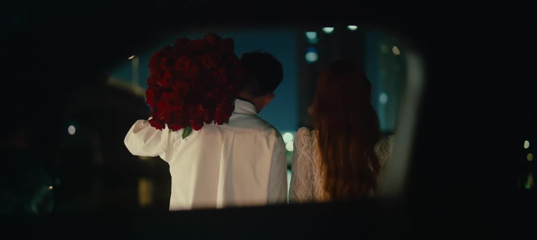 A romantic couple sits together in a car at night, viewed from behind, with the man holding a large bouquet of red roses