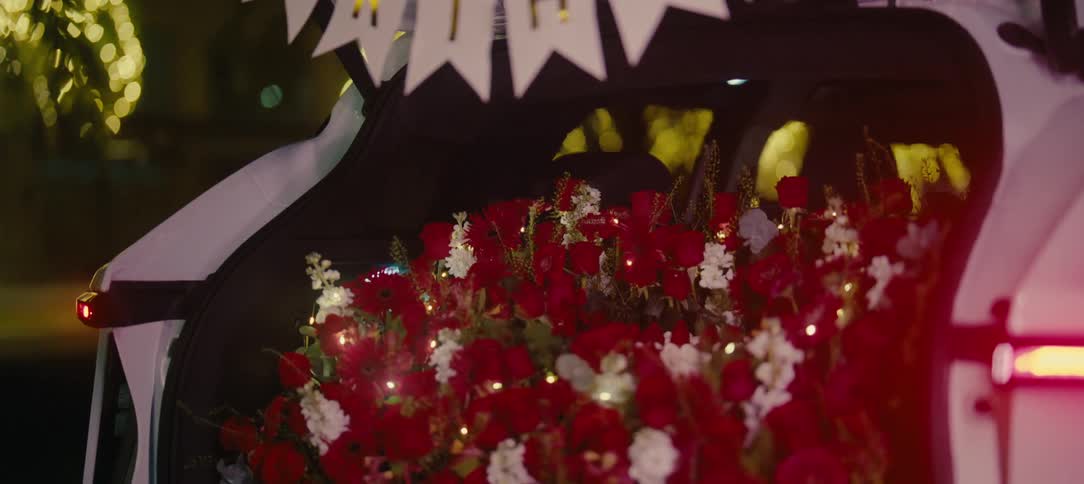 A romantic birthday surprise with a car trunk filled with a bouquet of red flowers, fairy lights, and a celebratory banner at night