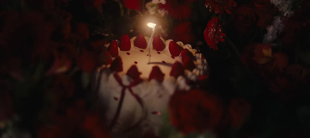 A single lit candle flickers on a strawberry birthday cake that is surrounded by red roses and flowers