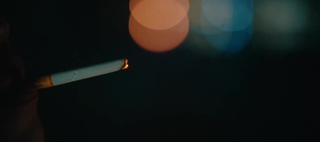 A close-up shot in slow motion of a hand lighting a cigarette with a torch lighter against a backdrop of city lights at night