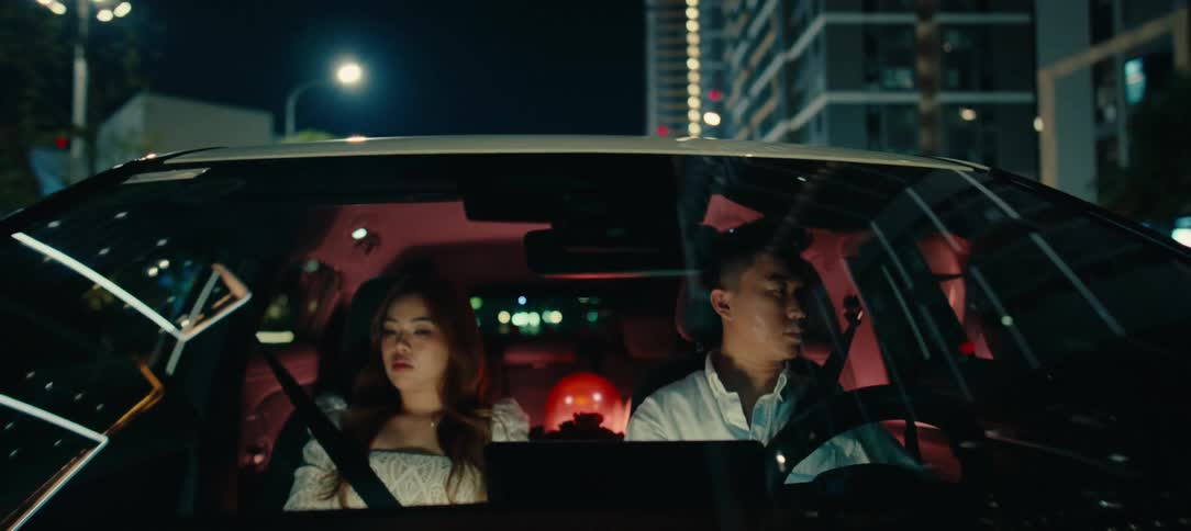 A somber young Asian couple drives through the city at night in silence, suggesting relationship problems