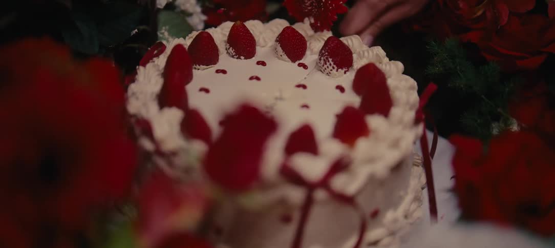 A close-up shot of a hand cutting a slice of a decorative strawberry and cream cake surrounded by red flowers
