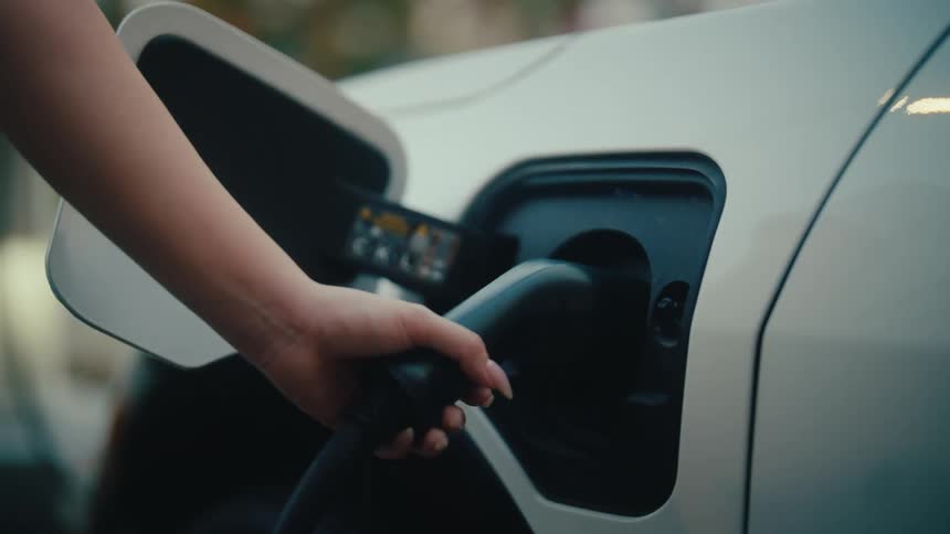 Close up shot of a woman's hand holding a charging plug connected to a modern white electric car