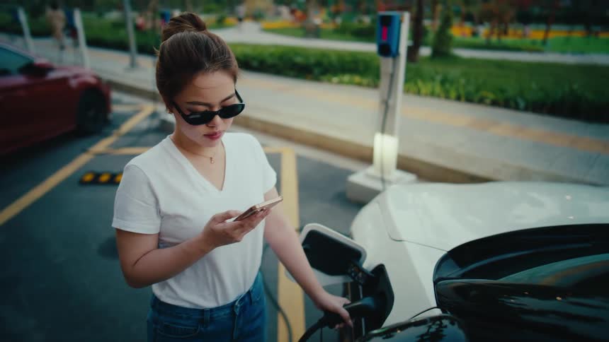 A fashionable young Asian woman uses her smartphone while waiting for her electric vehicle to charge at a public station