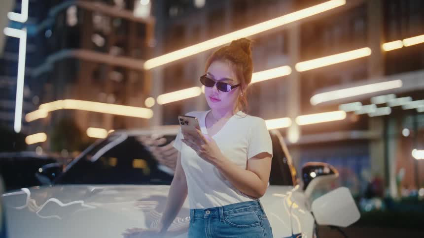 A stylish woman in sunglasses uses her smartphone while her electric vehicle charges at a brightly lit urban station at night