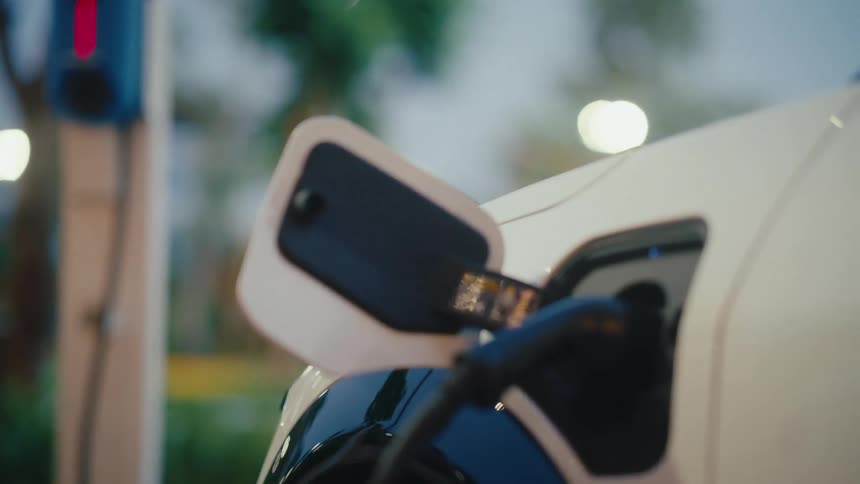 Focus shifts from an EV charging station to the charging port of a white electric car as it refuels its battery