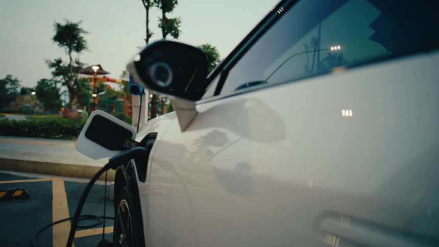A sleek white electric car charges at an outdoor station, with a slow dolly shot revealing the charging port at dusk