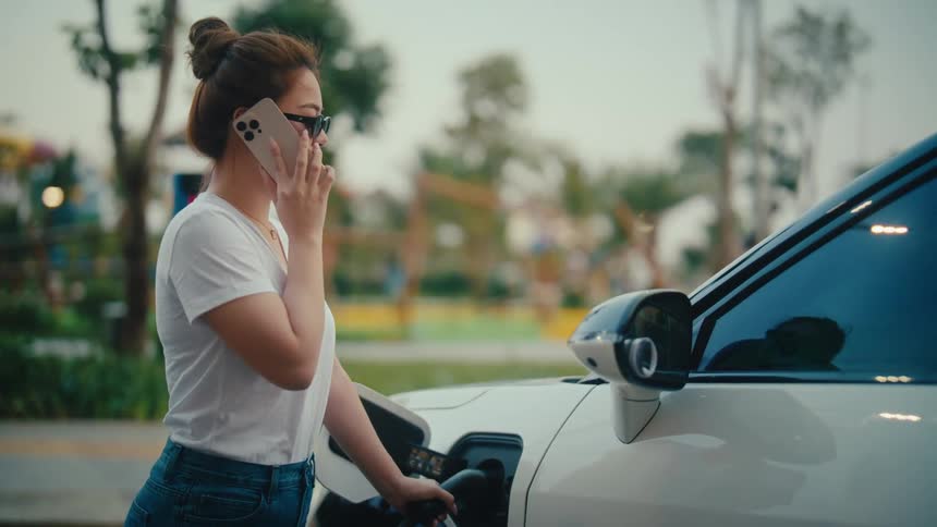 A stylish woman wearing sunglasses talks on her smartphone while her electric vehicle charges at a station