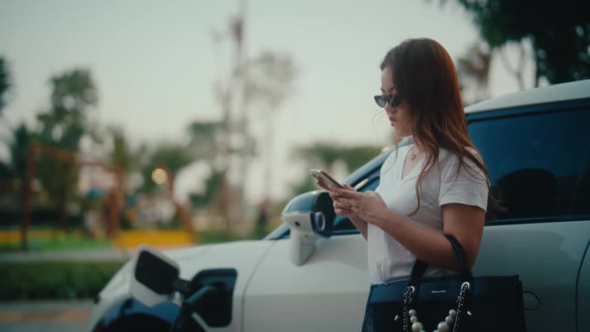 A stylish woman scrolls on her smartphone while leaning against her white electric car as it charges at an outdoor station