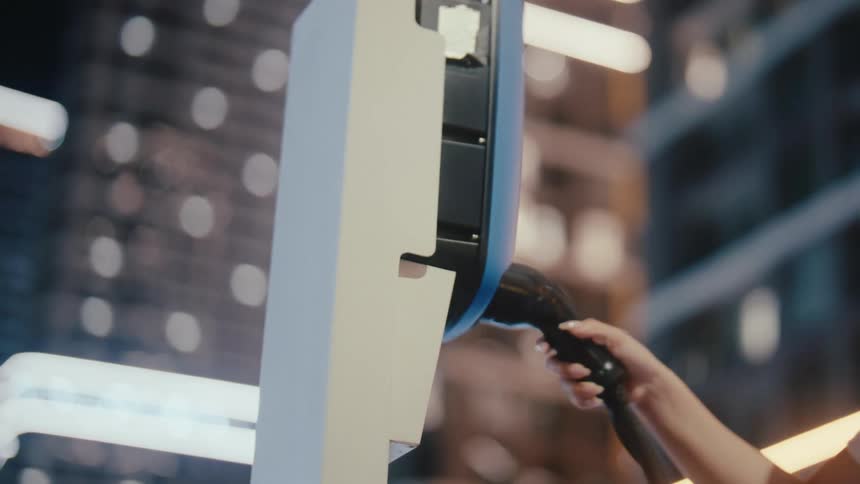 A hand slowly plugs a power cable into an electric vehicle charging station at night, with a modern cityscape blurred in the background