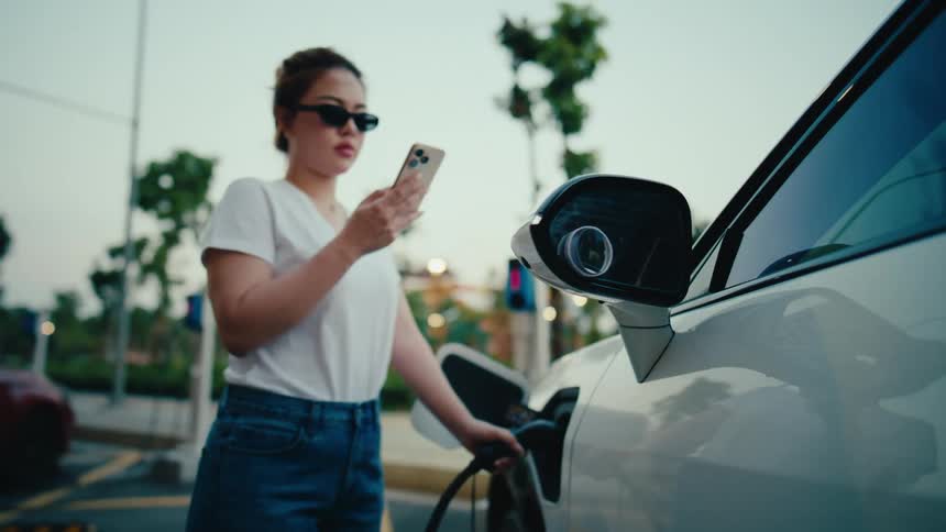 A slow zoom out reveals a stylish woman using her smartphone as her white electric vehicle charges at an outdoor public station
