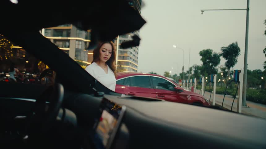 A woman plugs her red electric car into a charging station in a modern city at dusk, as seen from the interior of another vehicle