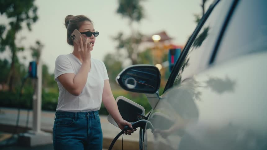 A stylish young Asian woman talks on her smartphone while her electric car charges at an outdoor station