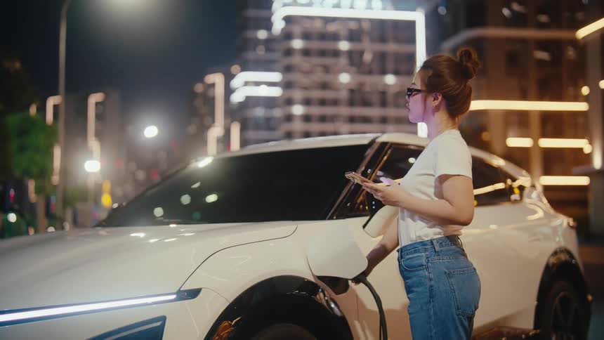 Stylish young woman scrolls on her phone while charging her futuristic electric car on a bustling city street at night