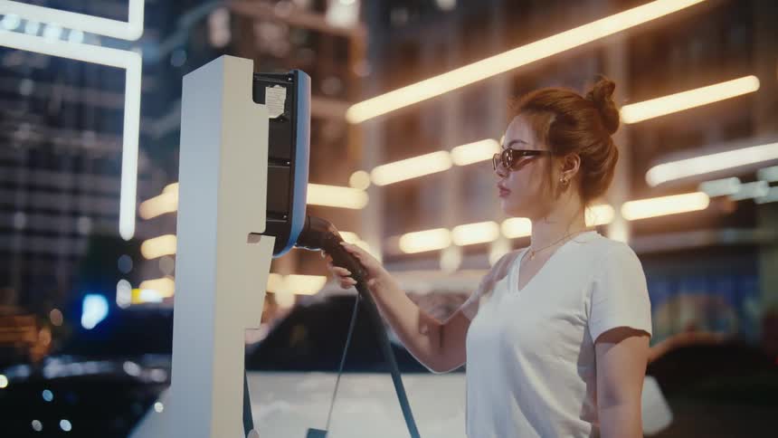 A young woman in sunglasses connects a charging cable to a public EV charging station in a modern city at night