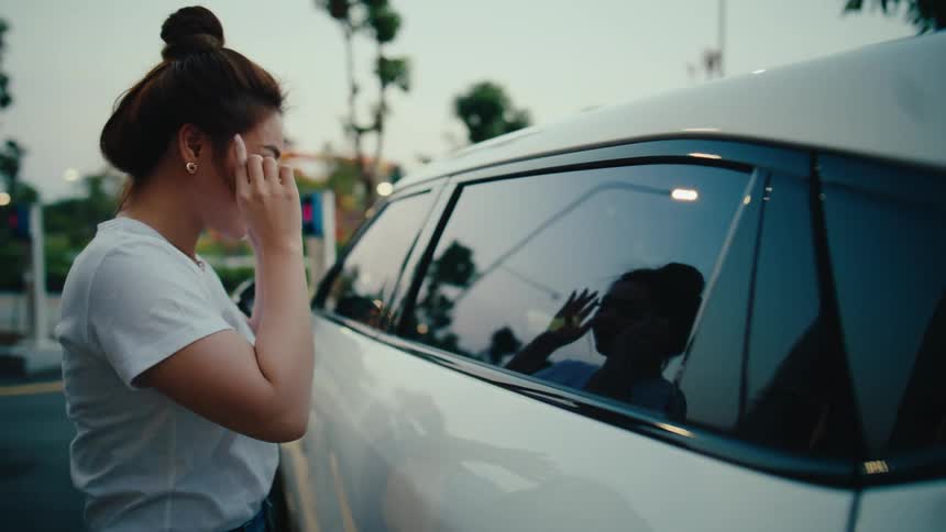 A young woman uses the reflection in a car window to tie her hair up while her friend is visible inside