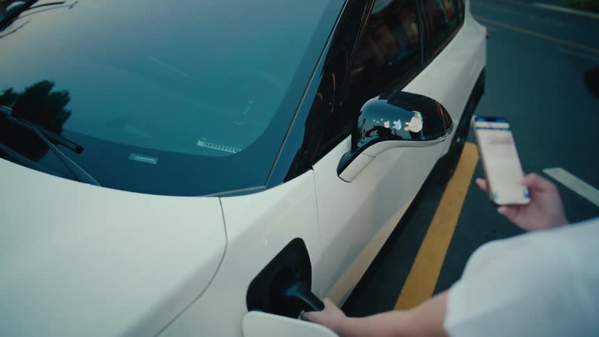 An over-the-shoulder view of a person using a smartphone app while their modern white electric car is charging