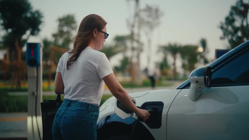 A woman plugs a charger into her white electric car at a public charging station with a park in the background