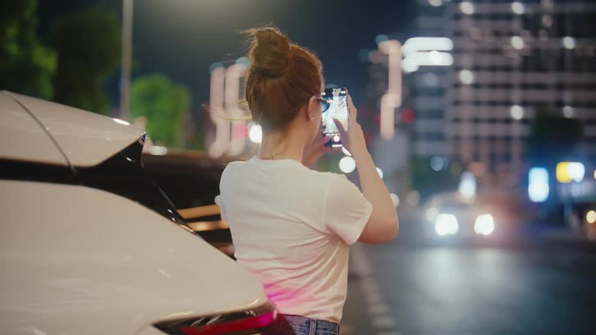 A young woman with her hair in a bun stands on a busy city street at night, capturing the scene with her smartphone