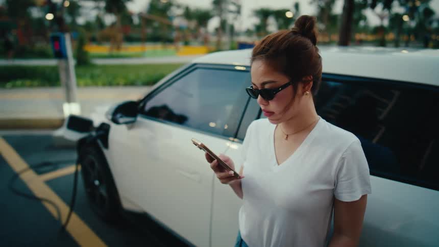 A woman wearing sunglasses uses her smartphone while leaning against her white electric vehicle at a charging station